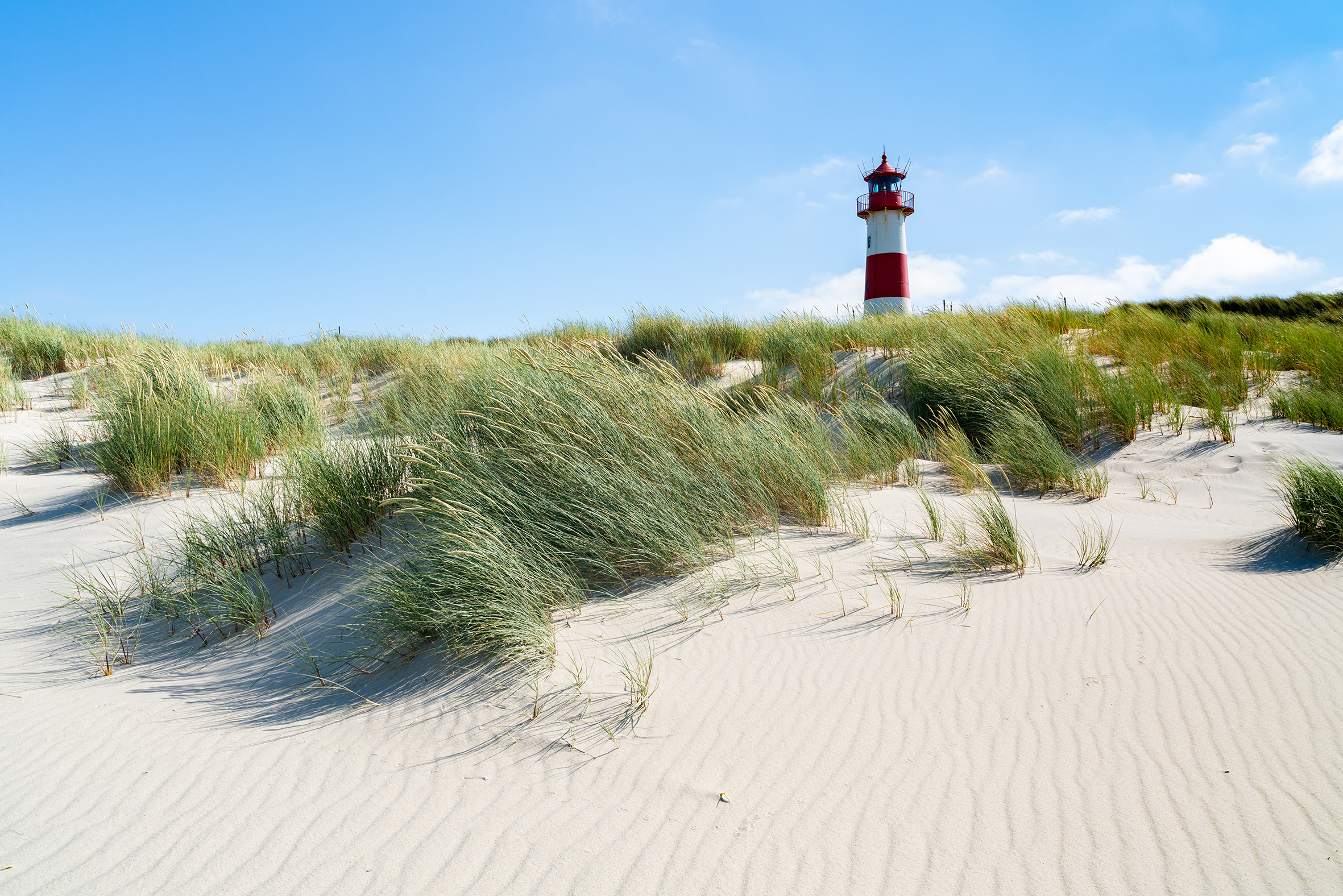 Roter und weißer Leuchtturm hinter sandigen Dünen mit Dünengras unter blauem Himmel mit wenigen Wolken.